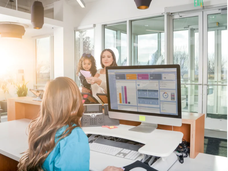 Receptionist di un salone di parrucchieri che accoglie una cliente con la sua bambina, utilizzando un computer con software gestionale per un'esperienza organizzata e professionale.